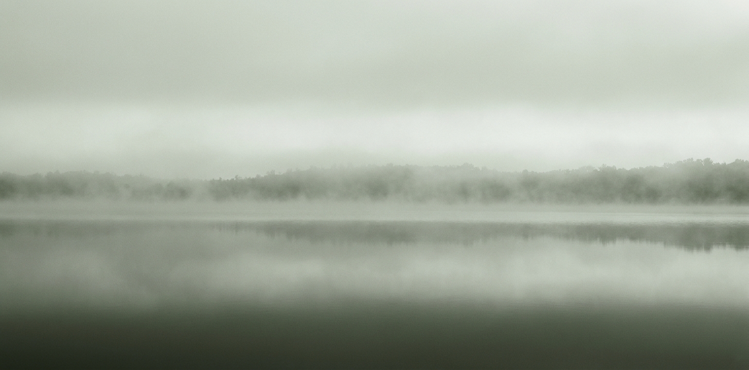 A misty Michigan lake at first light — water, fog, and sky stacked in soft sage-green bands